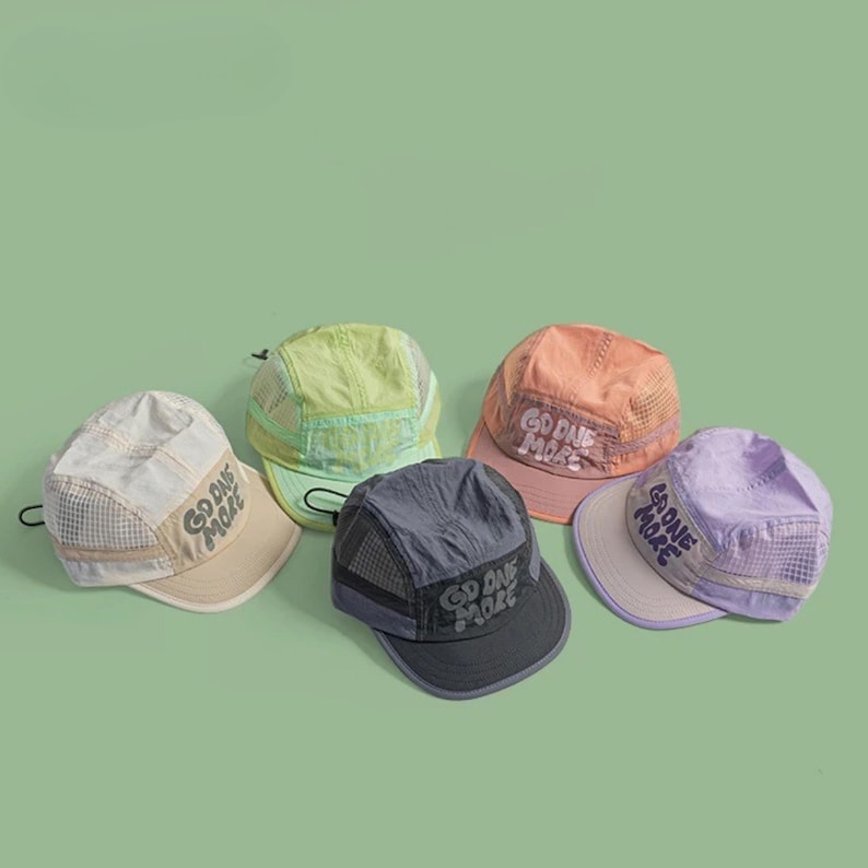May include: Five five-panel caps in beige, lime green, peach, dark gray, and lavender. Each cap features mesh side panels, a flat brim, an adjustable back strap, and the text "GO ONE MORE" embroidered in a stylized font on the front panel.