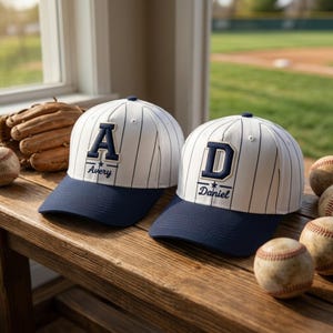 May include: Two white baseball caps with thin navy blue pinstripes and solid navy blue brims. Each cap features a large navy blue letter patch, with the name Avery on one and Daniel on the other, centered below the letter with a small star.