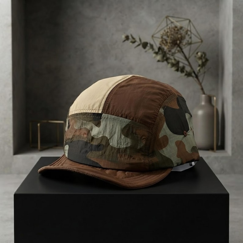 May include: A five-panel cap featuring a colour-blocked design with a beige top panel, a dark brown side panel, and a camouflage pattern band in shades of green, brown, and black. The cap includes a solid dark brown brim with visible stitching.
