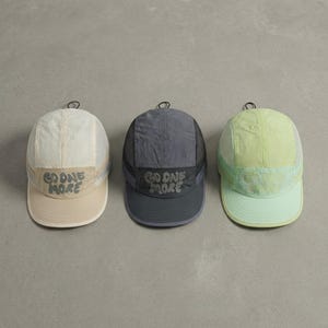 May include: Three five-panel caps in cream, charcoal gray, and light green. Each cap features mesh side panels, a flat brim, and the text "GO ONE MORE" embroidered in a stylized font on the front panel. The caps have a small loop at the top.