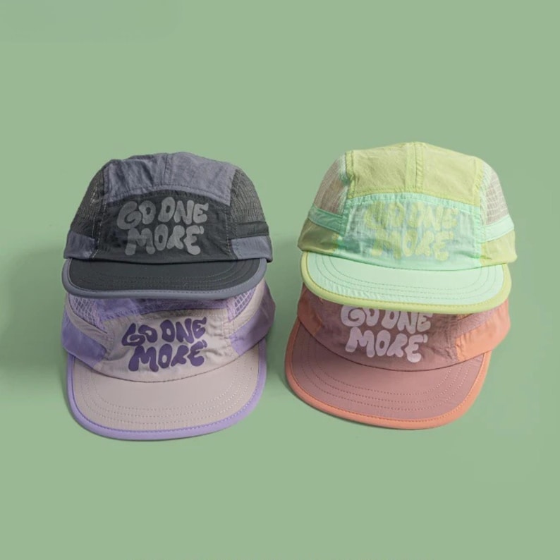 May include: Four five-panel caps featuring mesh side panels and the text GO ONE MORE printed on the front. The hats are colored in charcoal gray, light purple, pale yellow, and dusty rose, each with a matching brim and adjustable strap.