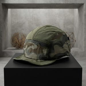 May include: A five-panel cap featuring a camouflage pattern in shades of olive green, brown, and black. The hat is constructed from a lightweight, crinkled fabric and includes a flat brim and a structured crown with a side panel detail.