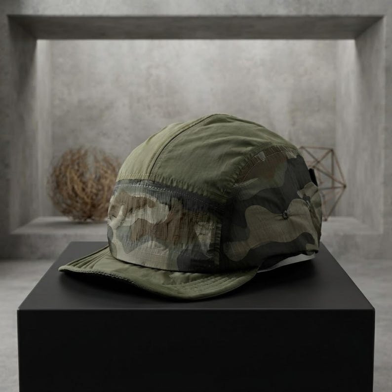 May include: A five-panel cap featuring a camouflage pattern in shades of olive green, brown, and black. The hat is constructed from a lightweight, crinkled fabric and includes a flat brim and a structured crown with a side panel detail.