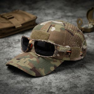 May include: A camouflage baseball cap with a perforated side panel and a rectangular hook-and-loop patch on the front. Resting on the brim are camouflage sunglasses with dark lenses and orange accents on the inner frame arms.