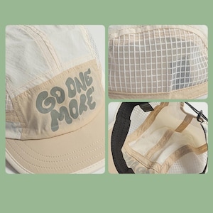 May include: A beige and off-white five-panel cap featuring a mesh side panel and the text GO ONE MORE in a dark grey font on the front panel. The cap includes a solid beige brim and an adjustable black cord strap on the interior back.