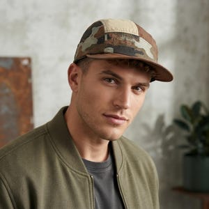 May include: A five-panel cap featuring a multi-coloured camouflage pattern in shades of brown, green, and tan. The hat has a structured crown with a solid tan top panel and a flat, brown brim. The design includes stitched seams and a fabric construction.