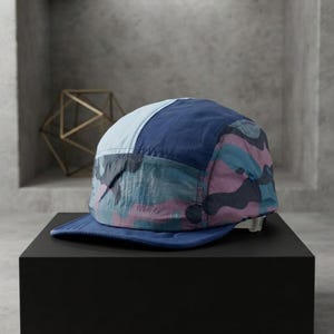 May include: A five-panel cap featuring a colour-blocked design with a light blue front panel, a dark blue side panel, and a camouflage pattern in shades of pink, blue, and dark grey. The cap has a solid dark blue brim and an adjustable back strap.