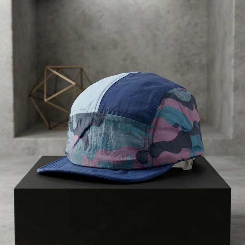 May include: A five-panel cap featuring a colour-blocked design with a light blue front panel, a dark blue side panel, and a camouflage pattern in shades of pink, blue, and dark grey. The cap has a solid dark blue brim and an adjustable back strap.