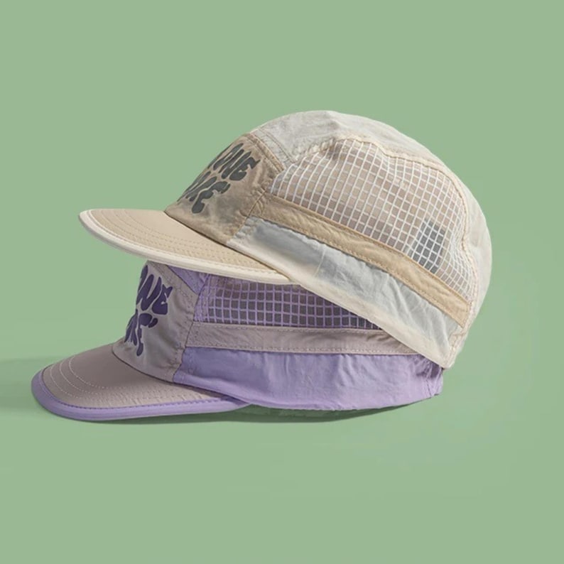 May include: Two stacked five-panel caps featuring mesh side panels and a flat brim. The top cap is beige with dark text, while the bottom cap is lavender with dark purple text. Both hats have a structured design with visible stitching and fabric panels.