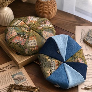 May include: Two berets are displayed. One is crafted from a green fabric featuring a repeating woven pattern of houses. The second beret is constructed from alternating panels of dark blue denim, light blue denim, and the same house-patterned fabric.