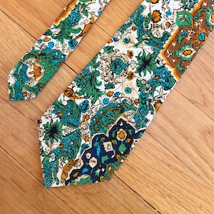 May include: A necktie featuring a complex, multi-colored pattern with shades of teal, forest green, mustard yellow, and dark blue on a white background. The design includes intricate floral and paisley-inspired motifs arranged in geometric sections.