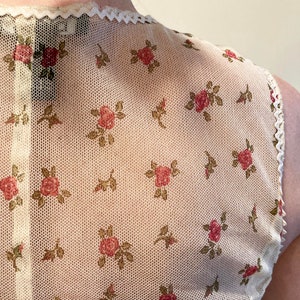 May include: A white mesh top with a floral pattern of small pink roses and green leaves. The top has a scalloped neckline and a white lace trim.