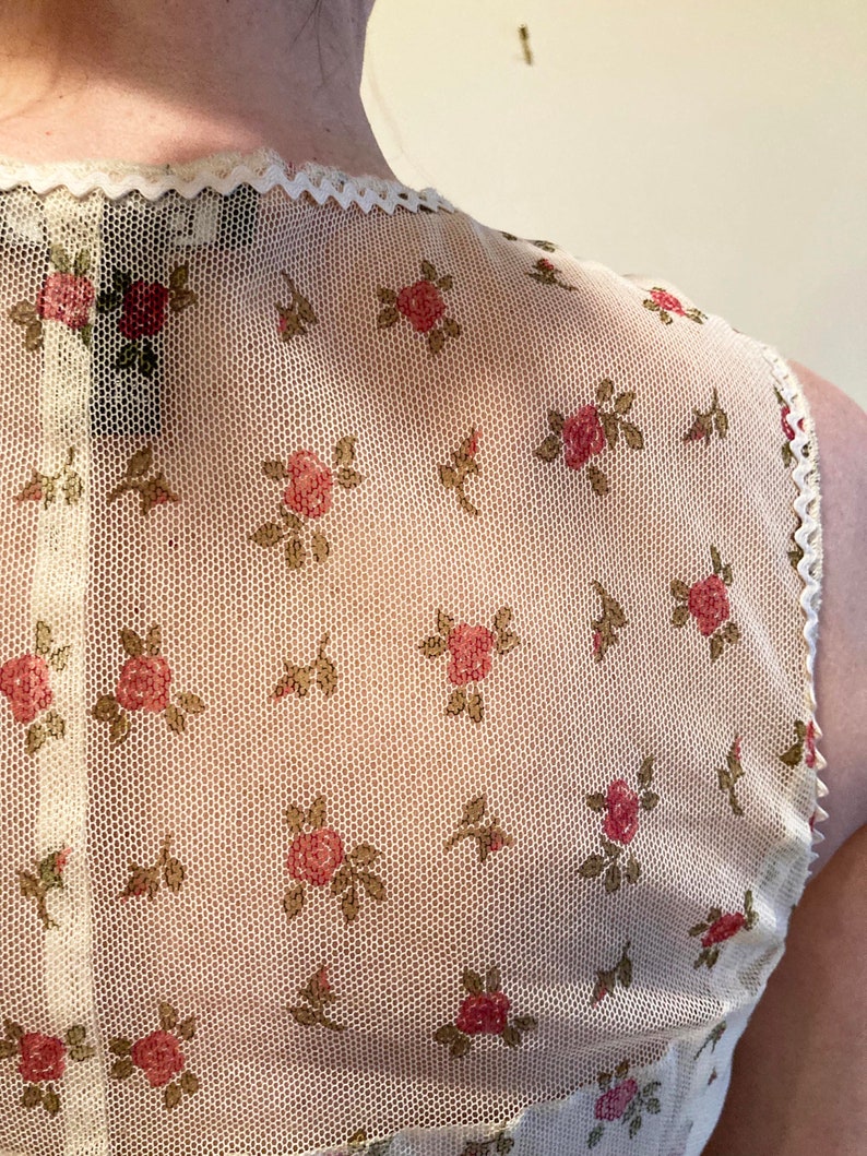 May include: A white mesh top with a floral pattern of small pink roses and green leaves. The top has a scalloped neckline and a white lace trim.