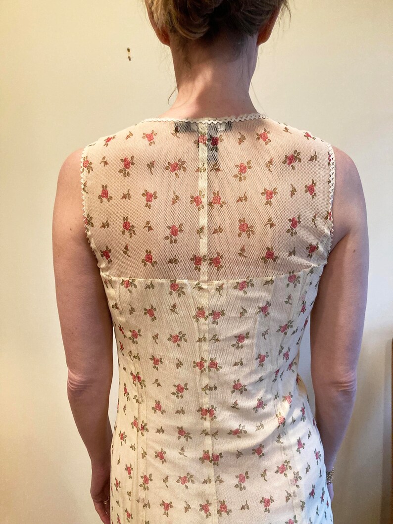 May include: A sleeveless white dress with a floral pattern and a sheer overlay. The dress has a fitted bodice and a flowing skirt.