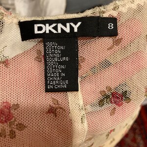 May include: A white DKNY brand tag with black text and a size 8 label. The tag is attached to a white mesh fabric with a floral pattern. The text on the tag reads "100% COTTON/COTON LINING/DOUBLURE: 100% COTTON/COTON MADE IN CHINA/FABRIQUE EN CHINE"
