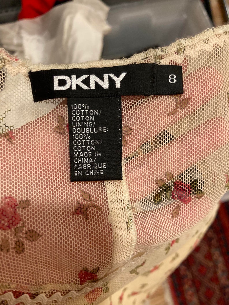 May include: A white DKNY brand tag with black text and a size 8 label. The tag is attached to a white mesh fabric with a floral pattern. The text on the tag reads "100% COTTON/COTON LINING/DOUBLURE: 100% COTTON/COTON MADE IN CHINA/FABRIQUE EN CHINE"