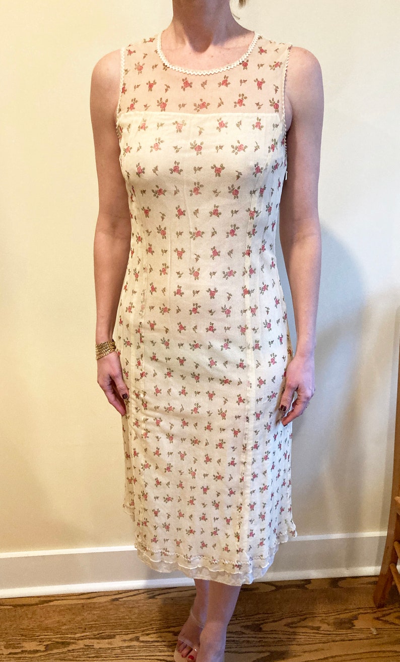 May include: A white sleeveless midi dress with a floral print of small pink roses. The dress has a sheer overlay at the neckline and a lace trim at the hem.