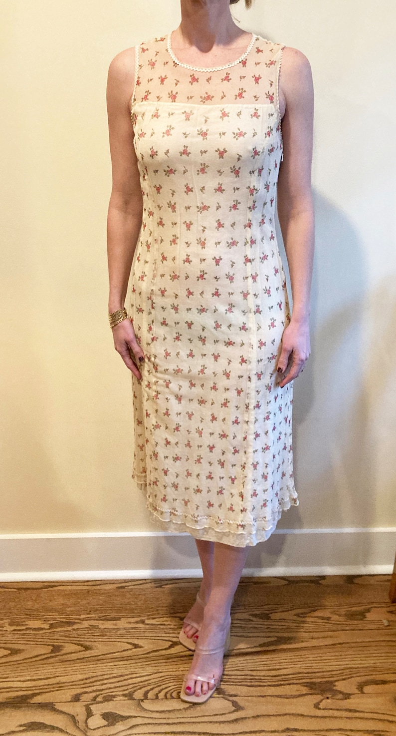 May include: A sleeveless midi dress in off-white features a sheer yoke with a delicate pearl trim and a bodice with a repeating pattern of small pink roses. The skirt flows to a scalloped hemline. The dress is made of a lightweight fabric, suitable for spring or summer.
