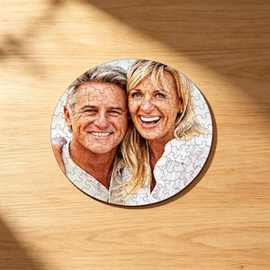 May include: A circular jigsaw puzzle featuring a photographic portrait of a smiling man and woman with blonde hair. The puzzle pieces are clearly visible over the image, which shows the two individuals from the chest up in bright lighting.