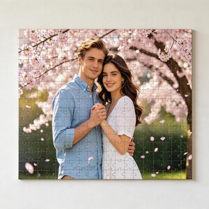 以下が含まれることがあります： 満開の桜の木の下に並んで立つ二人の写真がプリントされた正方形のジグソーパズル。左側の人物は水色のボタンダウンシャツを着用し、右側の人物は白いドレスを着用している。