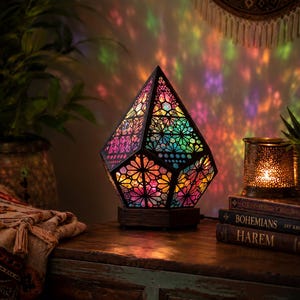 Può includere: Una lampada geometrica a forma di diamante con una cornice scura e pannelli traslucidi multicolori caratterizzati da intricati motivi a stella. La lampada emette una luce vivace e multicolore che proietta forme colorate sulla parete.