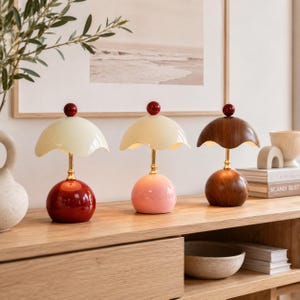 May include: Three mushroom-shaped table lamps with gold-tone stems and small red spheres on top. The first lamp has a cream shade and red base, the second has a cream shade and pink base, and the third has a wood-grain shade and wood-grain base.