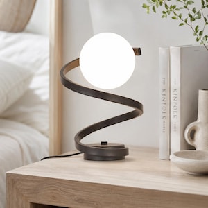 May include: A table lamp featuring a dark brown, spiral-shaped metal frame that supports a single, spherical white glass globe light. The lamp has a circular base with a small power switch and a black cord extending from the bottom of the fixture.