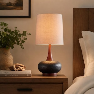 Può includere: Una lampada da tavolo con paralume cilindrico in tessuto beige, un collo sottile in legno e una base arrotondata e testurizzata color grigio scuro. La lampada emette una luce calda, illuminando lo spazio con un bagliore soffuso.