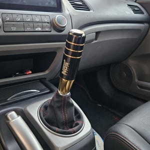 May include: A cylindrical black and gold gear shift knob featuring the text NOS RACING in white lettering, accented with horizontal gold rings. The knob is attached to a black leather shift boot with red stitching, installed in a vehicle interior.