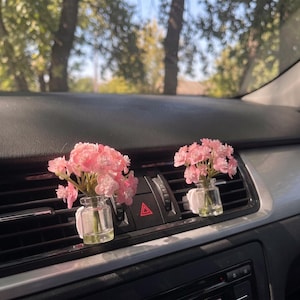 Può includere: Due piccole bottigliette in vetro trasparente contenenti fiori artificiali rosa, fissate alla bocchetta dell'aria di un'auto. Ogni bottiglietta presenta un mazzetto di delicati fiori rosa chiaro con steli verdi visibili, fissato tramite una clip bianca.