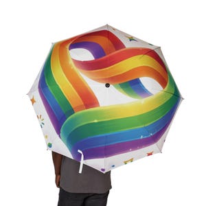 Könnte beinhalten: Ein weißer Regenschirm mit einem großen, wirbelnden Regenbogenband-Design, das sich über das Schirmdach zieht. Die farbenfrohe Grafik enthält leuchtende rote, orange, gelbe, grüne, blaue und violette Streifen, akzentuiert durch kleine Sterne und Punkte.