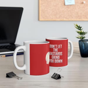 May include: A red ceramic mug featuring a white handle and interior. The exterior displays a graphic of a coffee bean and the white text "DON'T LET THE BASTARDS GRIND YOU DOWN" in a bold, distressed, sans-serif font on the side of the mug.