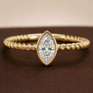 May include: A gold ring featuring a marquise-cut clear gemstone set in a bezel setting. The band of the ring has a twisted rope texture, creating a continuous metallic pattern that encircles the finger. The metal has a polished yellow gold finish.