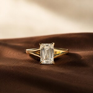 May include: A gold ring featuring a large, clear, emerald-cut gemstone held by four prongs. The band has a split-shank design that tapers towards the centre stone, showcasing a polished metallic finish and a faceted, rectangular centre stone.
