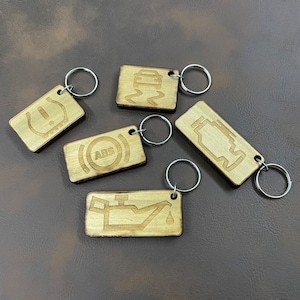 May include: Five wooden keychains with different car warning symbols. The symbols include a tire pressure warning, an ABS warning, an engine warning, a low oil warning, and a slippery road warning. Each keychain has a silver metal ring.
