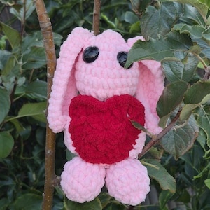 May include: A crocheted stuffed animal in the shape of a rabbit with light pink yarn, long ears, and two large black plastic eyes. The rabbit features a prominent, textured dark red heart shape attached to the centre of its torso area.