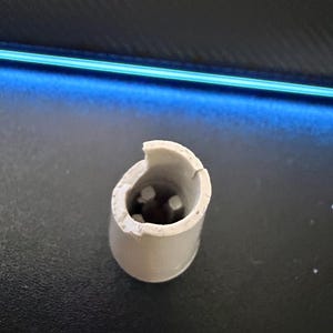 May include: A small, cylindrical, off-white plastic component with a hollow center and a jagged, broken top edge. The interior features three small, vertical, evenly spaced plastic prongs or connectors visible within the circular opening.