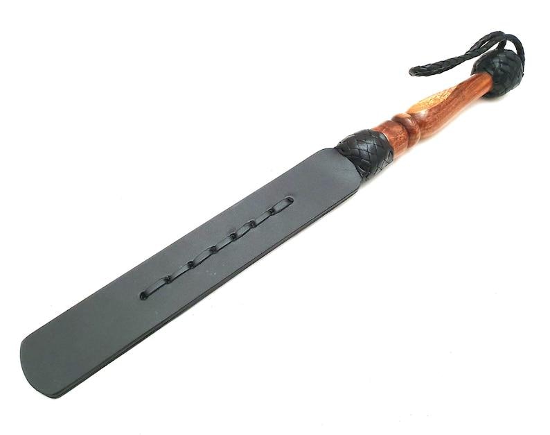 Handmade black leather paddle with wooden handle, crafted from premium genuine leather and polished hardwood. Durable leather craftsmanship accessory featuring braided leather accents, artisan handmade design, black leather paddle, leatherworking gift, handcrafted leather tool, rustic leather accessory, custom leather craftsmanship, traditional leather artisan product, premium quality leather item for collectors and handmade leather enthusiasts.
