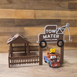 May include: A brown plastic playset featuring a small shed with a fenced enclosure and a sign that reads TOW MATER TOWING & SALVAGE. A small brown and blue toy tow truck with a yellow number 95 on top is positioned in front of the sign.