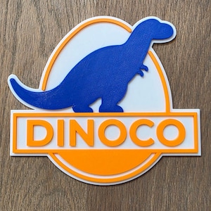 May include: A blue dinosaur silhouette within a white and orange oval, with the word "DINOCO" in orange block letters beneath. The design is set against a wooden background.
