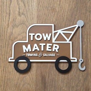 May include: A brown and white wooden sign shaped like a tow truck. The sign displays the words "TOW MATER - TOWING & SALVAGE" in white lettering. The truck features black wheels and a silver tow hook.