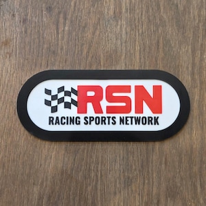 May include: An oval-shaped sign with a white background and a thick black border. It features a black and white checkered flag icon, the red letters RSN, and the text RACING SPORTS NETWORK in black capital letters centred below the acronym.