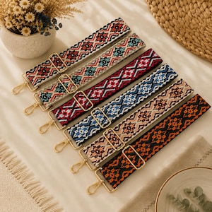 May include: A collection of six adjustable bag straps featuring intricate woven geometric patterns in various color combinations including red, teal, blue, and orange, each equipped with gold-toned metal swivel hooks and sliding buckles.
