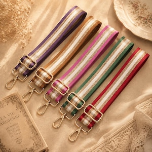 May include: Five adjustable bag straps in purple, brown, magenta, green, and red. Each strap features a central white stripe flanked by two shimmering gold glitter lines. The straps include gold-toned metal sliders and lobster claw clasps.