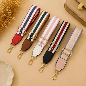 Könnte beinhalten: Fünf verstellbare Taschengurte mit gewebten Streifenmustern in Rot und Blau, Grün und Braun, Rosa und Weiß, Kastanienbraun und Schwarz sowie Hellblau und Beige. Jeder Gurt verfügt über goldfarbene Metallbeschläge und drehbare Karabinerhaken.