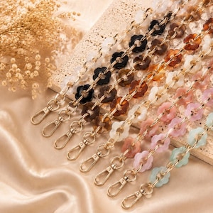 May include: A collection of bag straps made from interlocking acrylic flower-shaped links in various colors including black, brown, white, pink, and light blue, each featuring a gold-toned metal lobster clasp at the end for secure attachment.