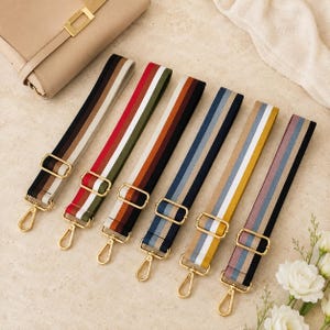 May include: Six adjustable bag straps made of woven fabric with various striped patterns in colors including beige, red, olive green, brown, navy blue, yellow, and grey. Each strap features gold-toned metal hardware, including sliding buckles and swivel lobster clasps.