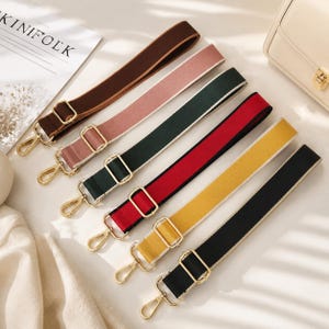 May include: A collection of six adjustable bag straps in brown, dusty pink, dark green, red, yellow, and black. Each strap features a solid color fabric band with white edges, gold-toned metal sliders for length adjustment, and gold-toned swivel hooks.