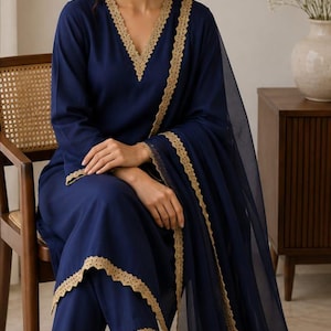 Op de afbeelding: Een diep marineblauwe driedelige set bestaande uit een tuniek met lange mouwen en een V-hals, een bijpassende broek en een transparante dupatta. Elk kledingstuk is afgewerkt met een geschulpte gouden kanten rand langs de zomen.