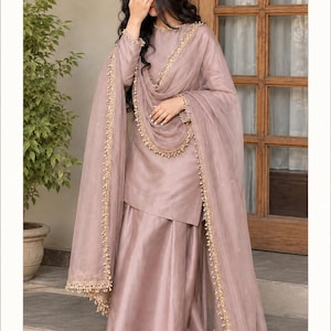 Könnte beinhalten: Ein malvenfarbenes Set aus einer langärmeligen Tunika und einer weiten Hose, kombiniert mit einem passenden, transparenten Dupatta. Das Ensemble ist mit zarten goldfarbenen Stickereien und kleinen perlenartigen Verzierungen an den Rändern versehen.